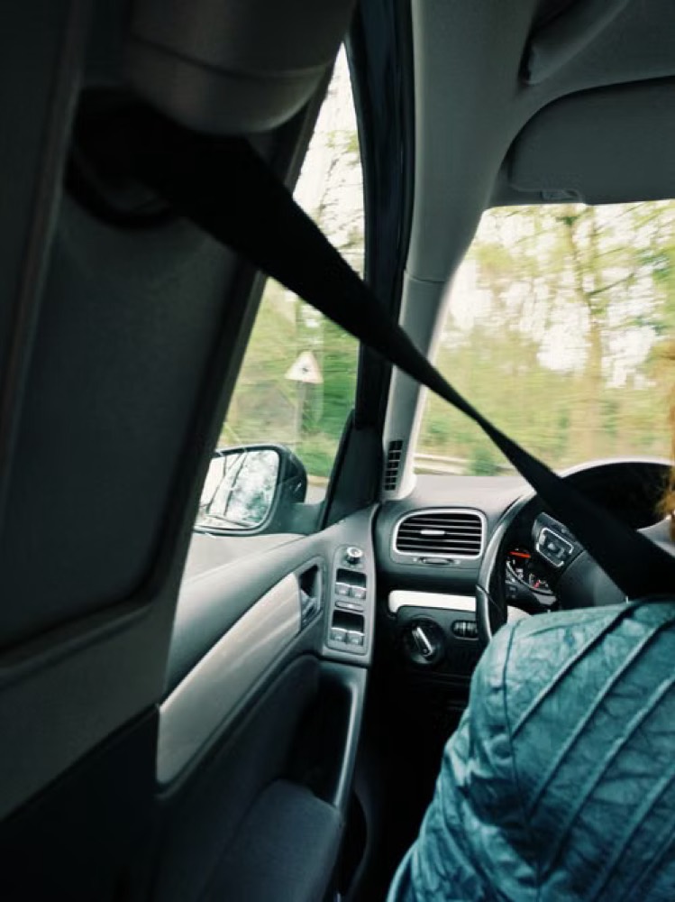 The interior of a van looking forward from behind the driver, with the seat belt visible across the seat and the road ahead through the windscreen.