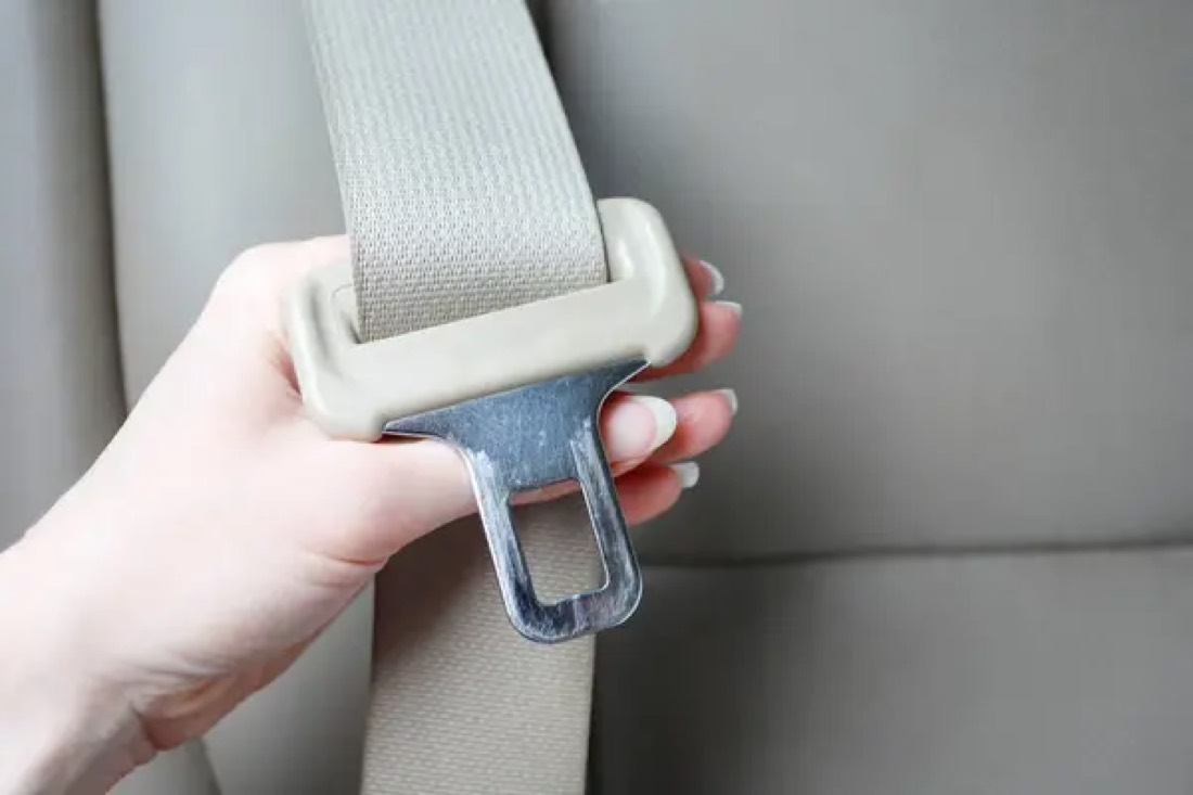A hand holding a pale seat belt tongue against a beige vehicle seat, with the latch aperture clearly visible.