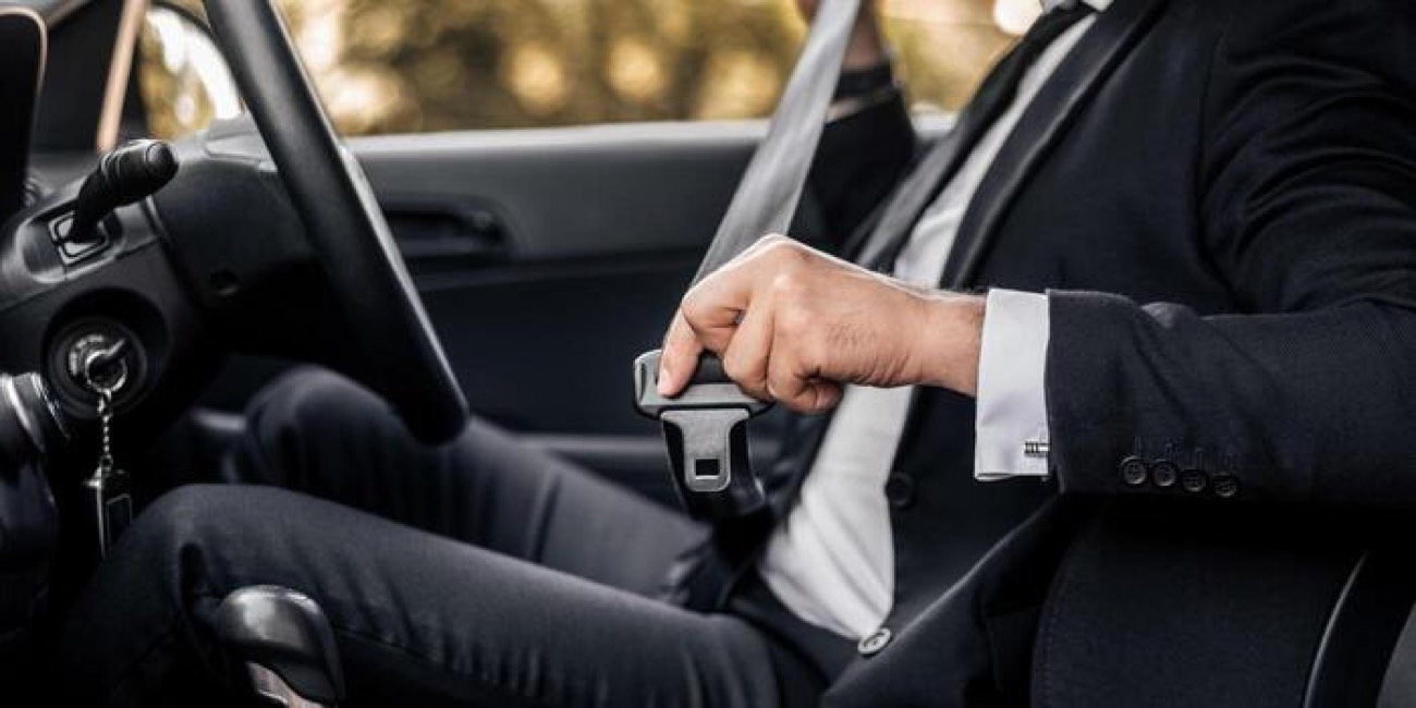 A person in a dark suit seated in a car, hand resting on the seat belt buckle beside the seat.