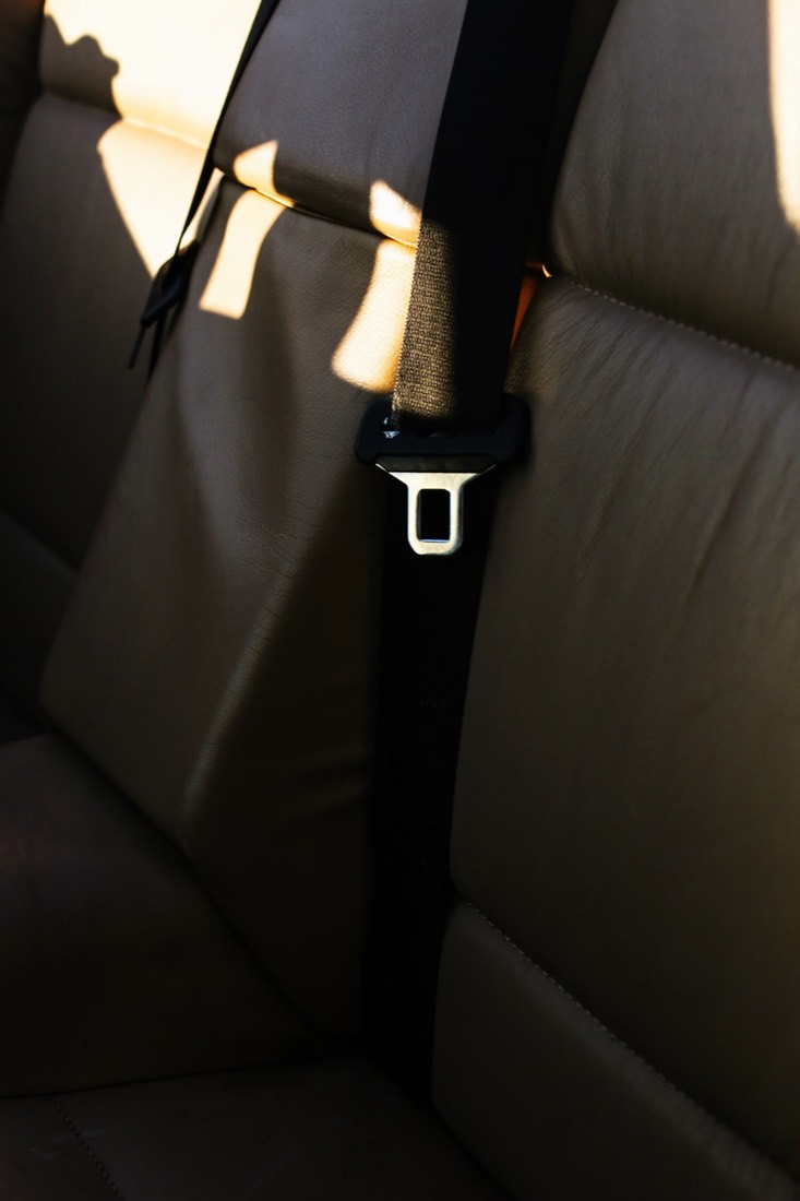 A black seat belt running diagonally across a pale leather rear bench seat, lit by low sunlight through a side window.