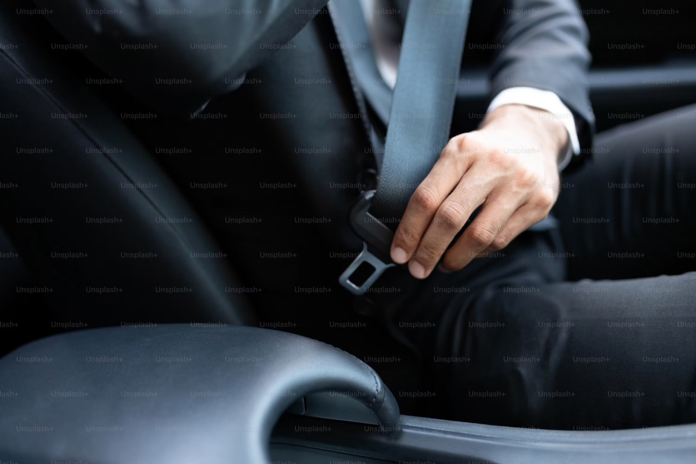 A person in a dark suit reaching for the seat belt buckle beside the seat, photographed close up inside a vehicle.